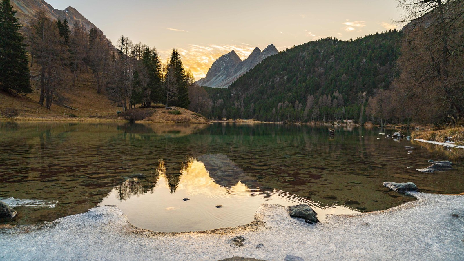 Bergsee mit spiegelnden Gipfeln und goldenem Licht am Horizont